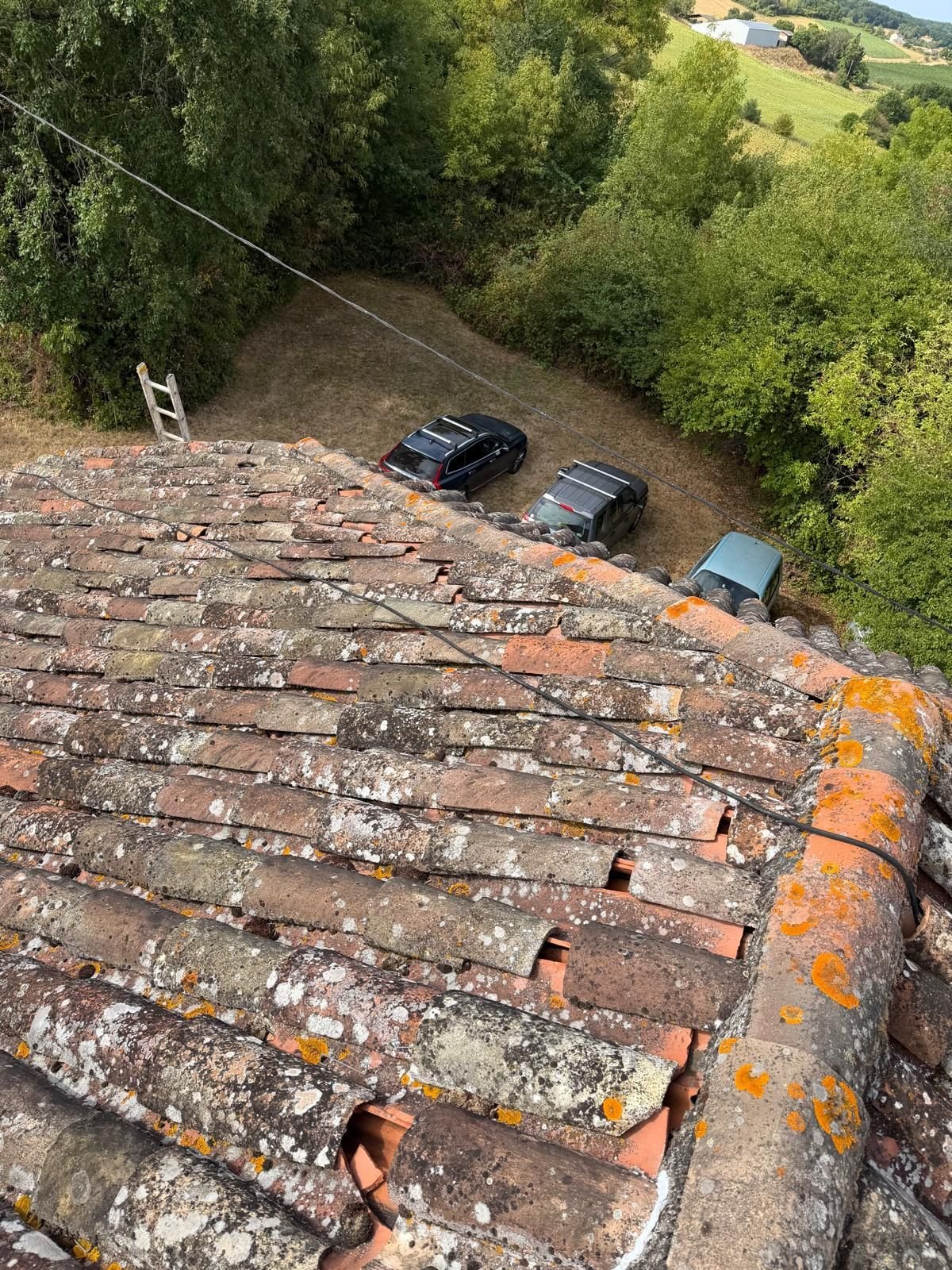 Vue plongeante sur un versant en tuile canal dégradé, tuiles cassées et manquantes, véhicules stationnés en contrebas.
