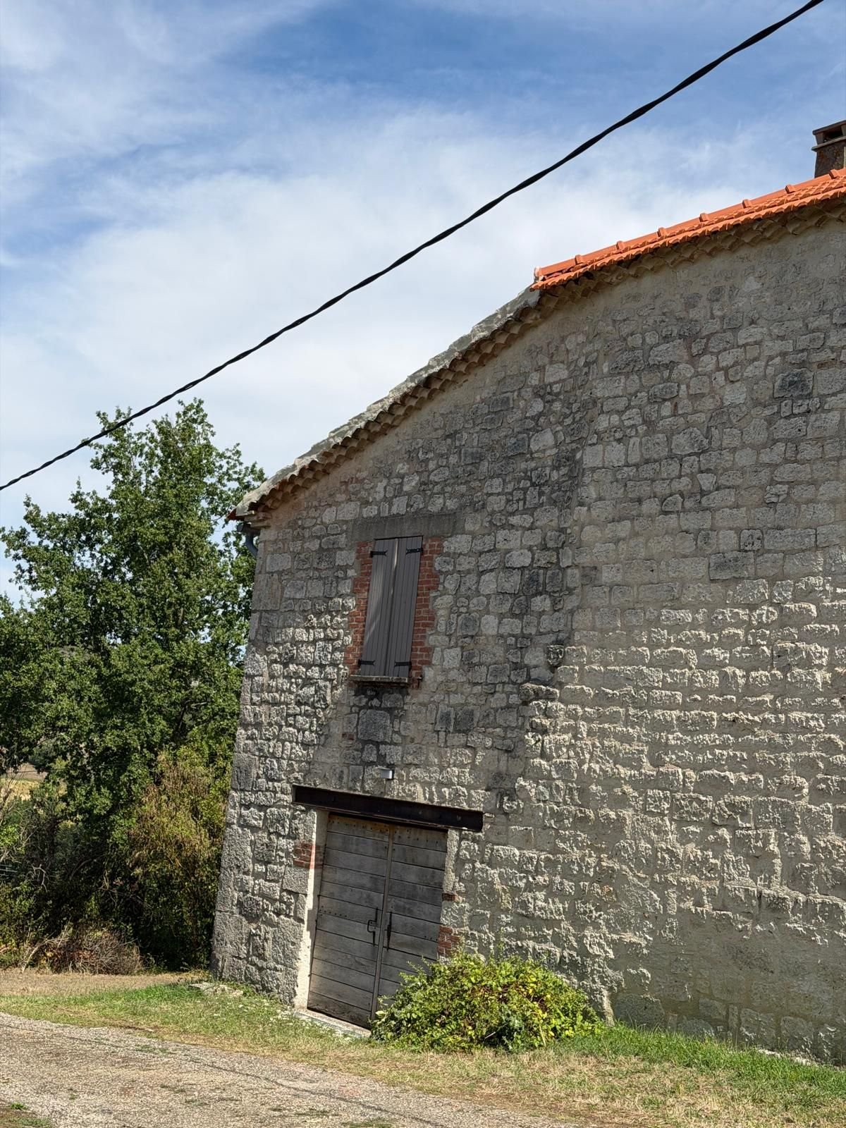 Pignon aveugle d'une grange en pierre, avec la toiture du corps principal visible à l'arrière-plan.