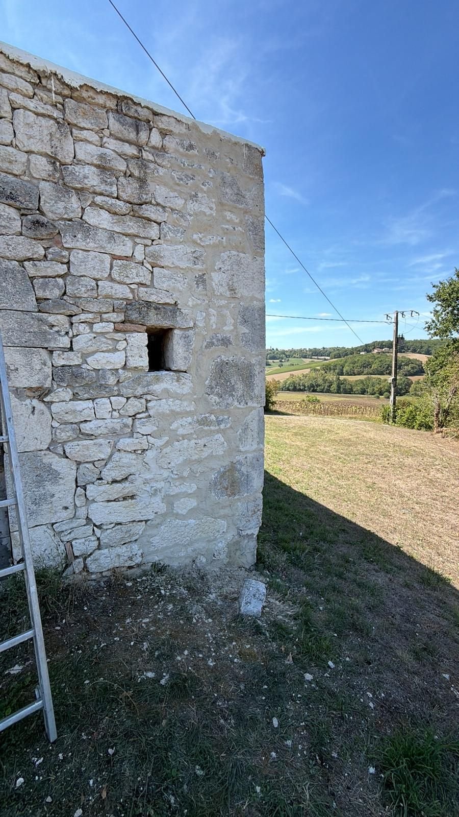 Pignon de la tour en pierre appareillée, échelle dressée contre le mur, paysage de coteaux au loin.